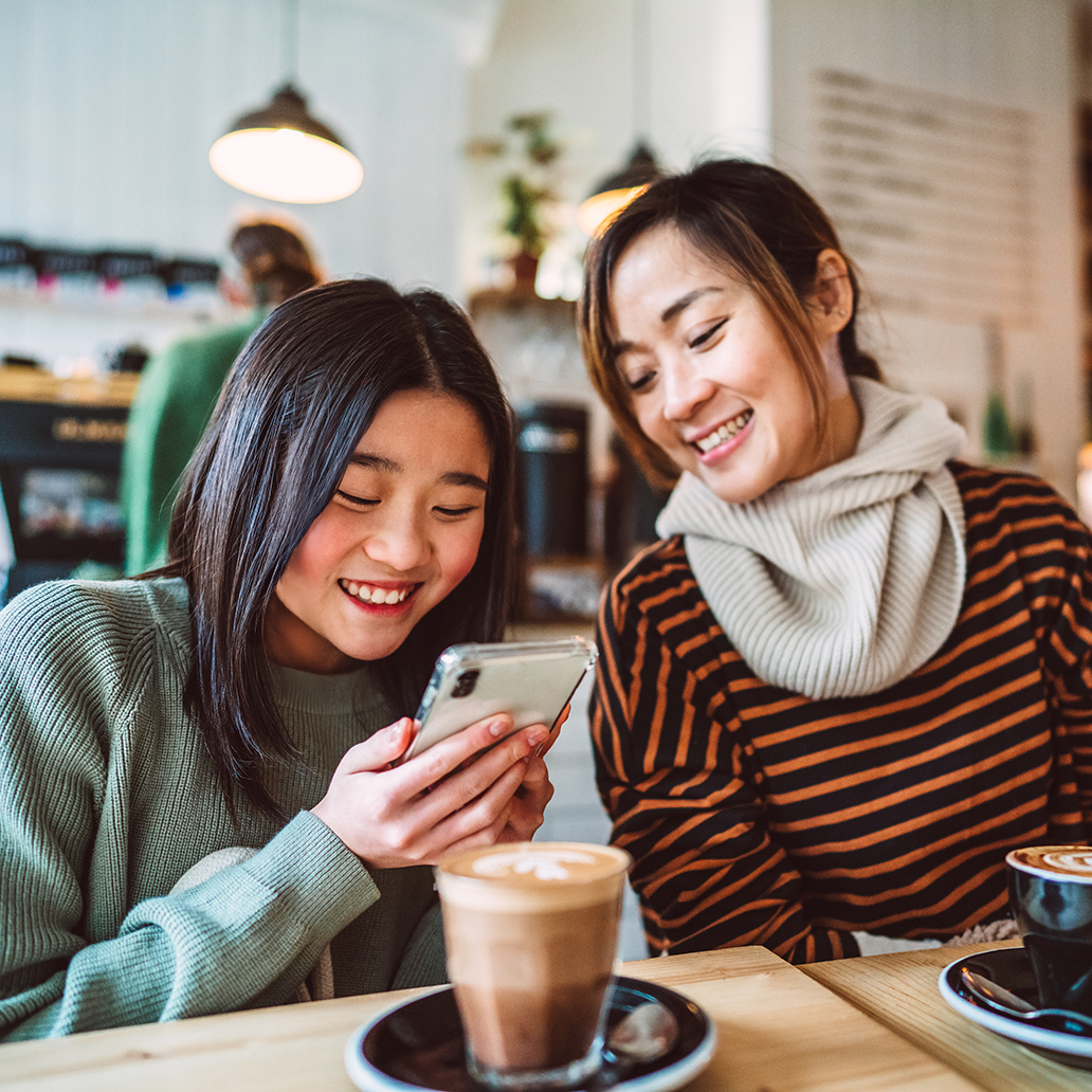Cheerful mother and teenage daughter using smart phone together while enjoying coffee break in a cafe