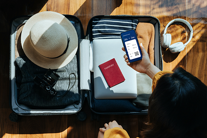Overhead view of Asian woman holding smartphone showing electronic flight ticket above an open suitcase with clothings, sun hat, camera, headphones, laptop and passport on wooden floor against sunlight. Traveller's accessories. Travel and vacation concept
