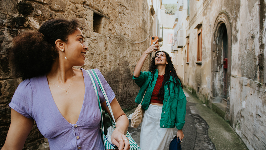 Two stylish woman walk down an alleyway. One takes a photo using her mobile phone.