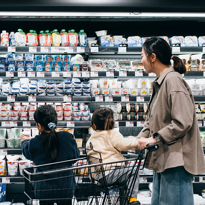 Young Asian mother and daughters shopping together joyfully in supermarket. They are standing in front of the display fridge with assorted dairy products. Healthy family eating lifestyle. Making healthier food choice for kids