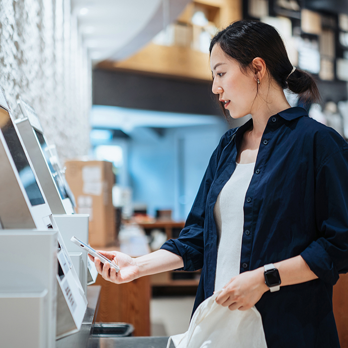 Young Asian woman with a reusable shopping bag, using contactless payment via smartphone to pay for her shopping at self-checkout kiosk in a store
